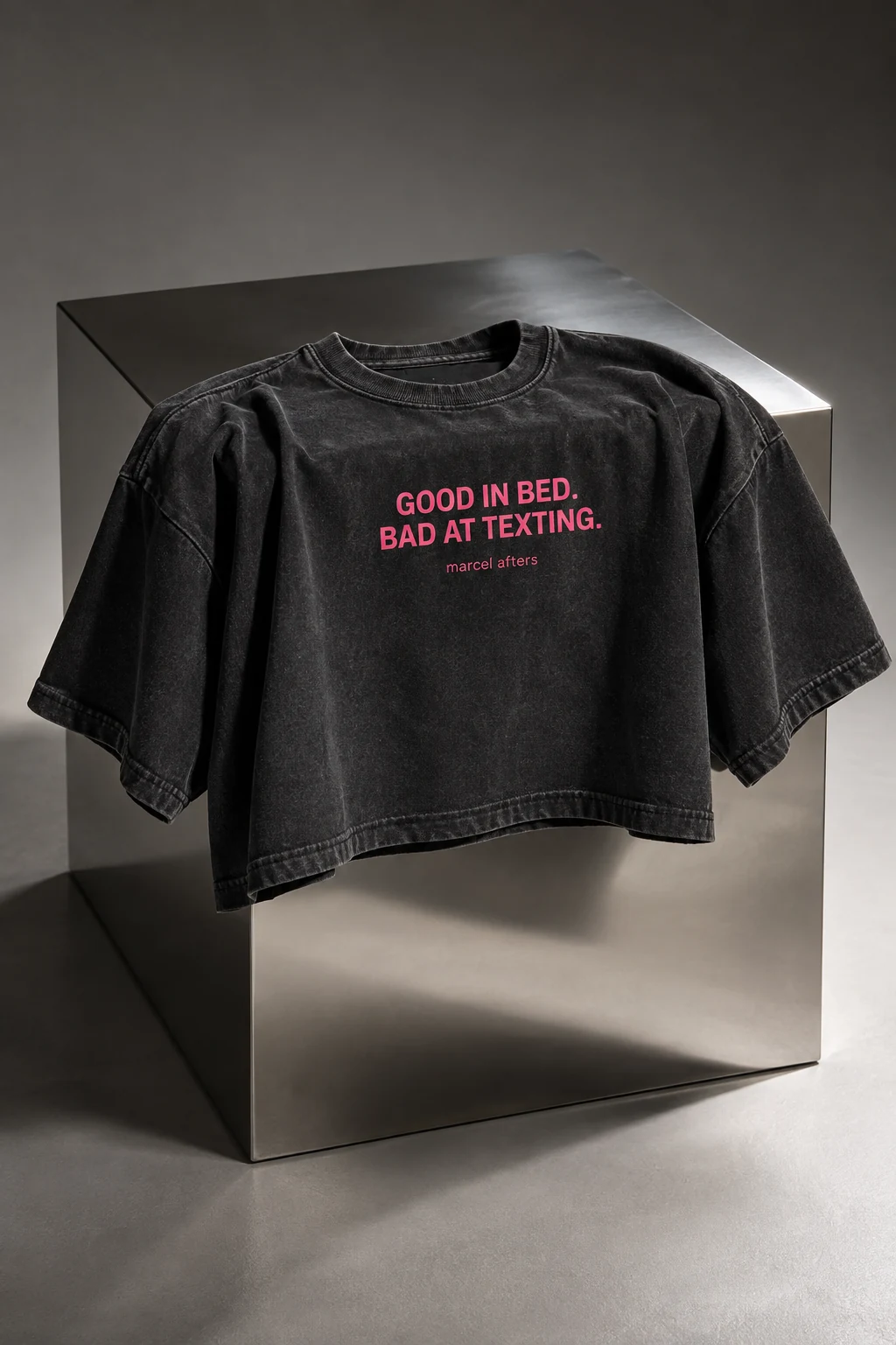 Charcoal Good in Bed. Bad at Texting. shirt concept displayed on a reflective block, with pink slogan and Marcel Afters signature visible.