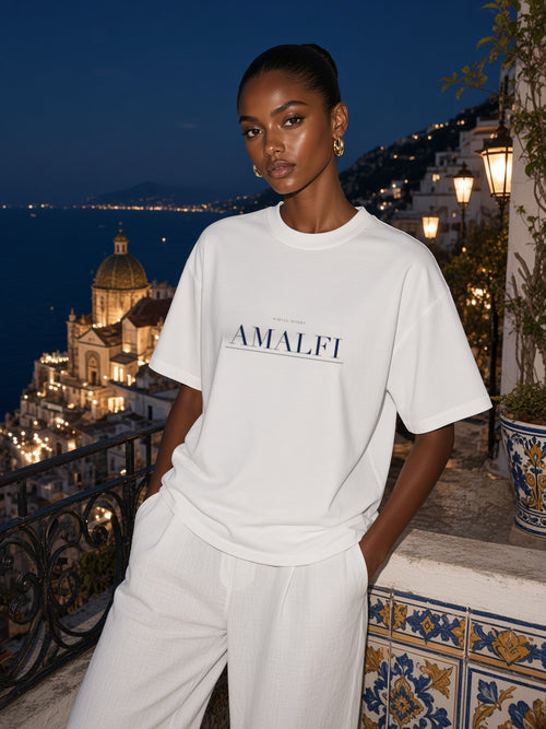 Woman in the Amalfi Heavyweight Drop Shoulder T-Shirt in White with the navy Amalfi city plate, on a tiled balcony above Amalfi at night