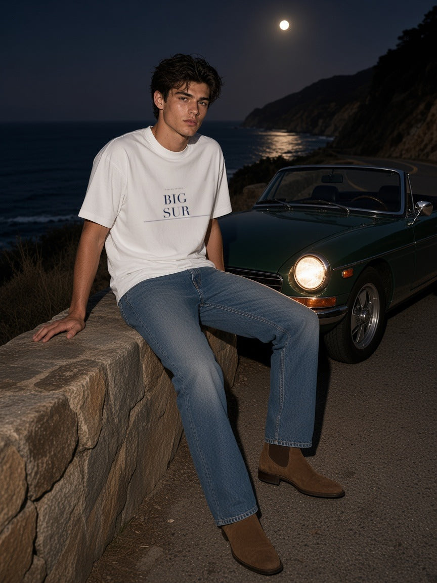 Man in the Big Sur Heavyweight Drop Shoulder T-Shirt in White with the navy Big Sur city plate, seated on a stone wall above the coast with a vintage car behind him in Big Sur at night