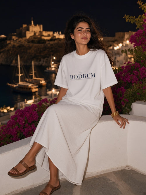 Woman in the Bodrum tee in White seated on a whitewashed terrace wall above Bodrum harbour at night