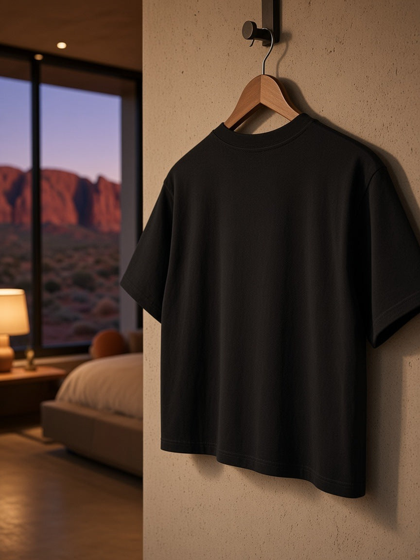 Boxy Drop Shoulder Crop T-Shirt in Black on a wooden hanger hooked on a pale plaster wardrobe in a Utah desert suite at night