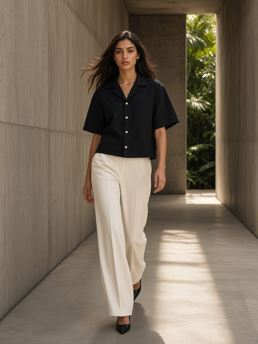Woman in the Boxy Heavyweight Short Sleeve Shirt in Black with ivory wide trousers, walking a stone corridor in afternoon light