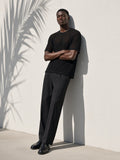 Man in the Boxy Mesh Crewneck T-Shirt in Black with charcoal trousers and loafers, leaning on a white wall in palm shadow