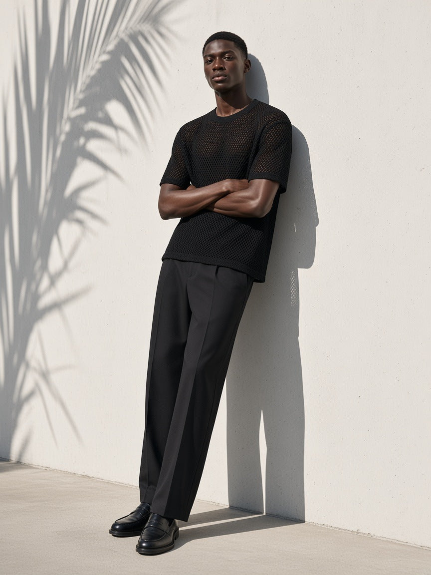 Man in the Marcel Afters Boxy Mesh Crewneck T-Shirt in Black with charcoal trousers and loafers, leaning on a white wall in palm shadow