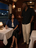 Two people in the Boxy Patch Pocket Short Sleeve Shirt in Navy Blue and Black, one seated at a window table and one standing mid-conversation, in a night train dining car