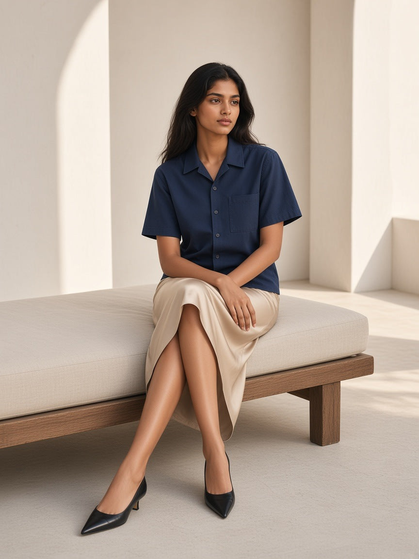 Woman in the Marcel Afters Boxy Patch Pocket Short Sleeve Shirt in Navy Blue with a sand skirt and pointed heels, seated on a daybed in a limestone room