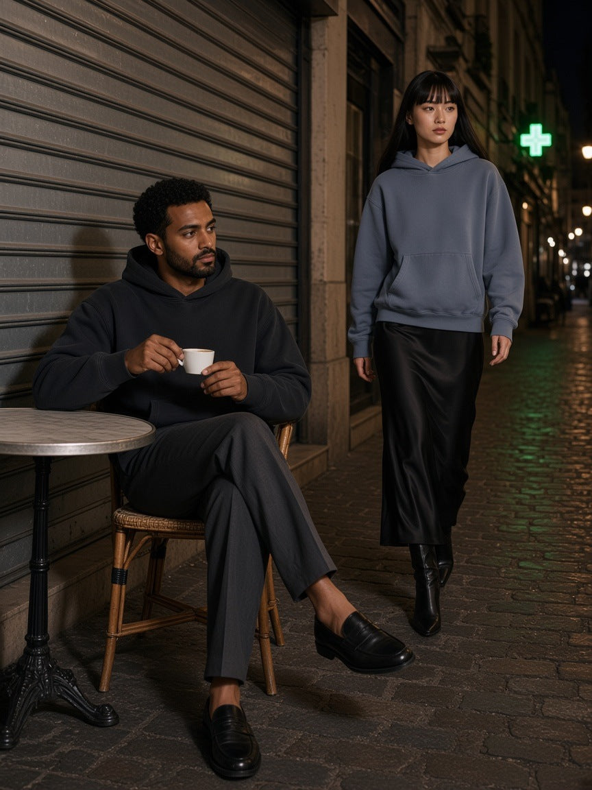 Two people in the Boxy Vintage Washed Hoodie in Black and Gray Blue, a man at a zinc cafe table and a woman walking past on Rue Saint-Denis in Paris at night