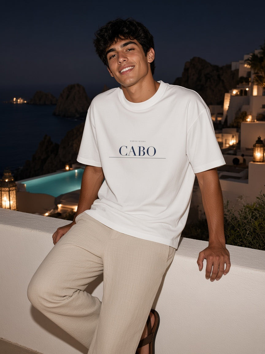 Man in the Cabo Heavyweight Drop Shoulder T-Shirt in White with the navy Cabo city plate, on a terrace above the marina lights in Cabo at night