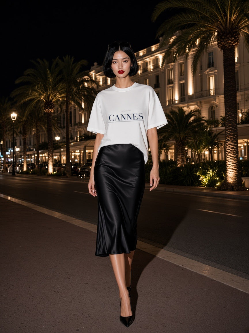 Woman in the Cannes Heavyweight Drop Shoulder T-Shirt in White with the navy Cannes city plate, walking the palm-lined seafront of Cannes at night