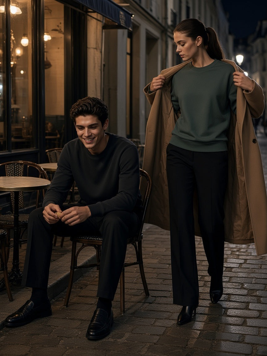 Two people in the Classic Cotton Sweatshirt in Classic Black and Olive Green, one seated at a café table and one pulling on a camel coat, in the Marais in Paris at night