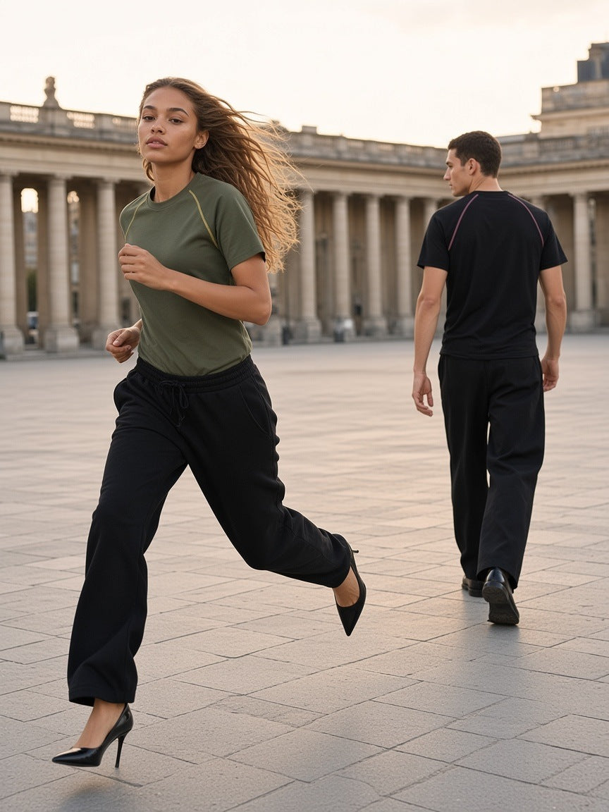 Two people in the Marcel Afters Contrast Stitch Raglan T-Shirt, her in Army Green running in heels and him in Black walking behind, across a stone colonnade square at dusk