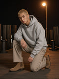 Man in the Contrast-Stitched Boxy Fleece Hoodie in Flower Gray with stone pleated trousers, kneeling on the Brooklyn Bridge Park pier at night