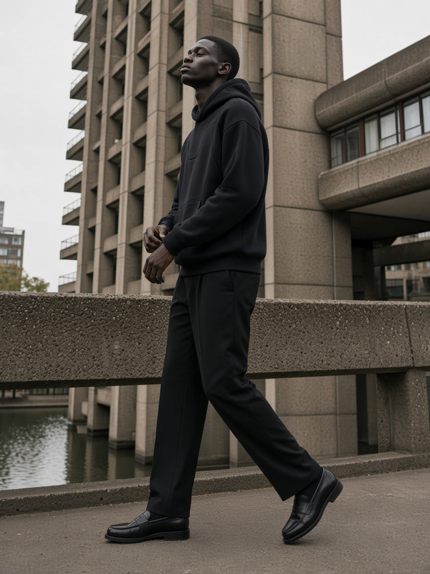 Man in the Curved Panel Raglan Hoodie in Black with black pleated trousers and loafers, walking a concrete walkway at the Barbican in London by day