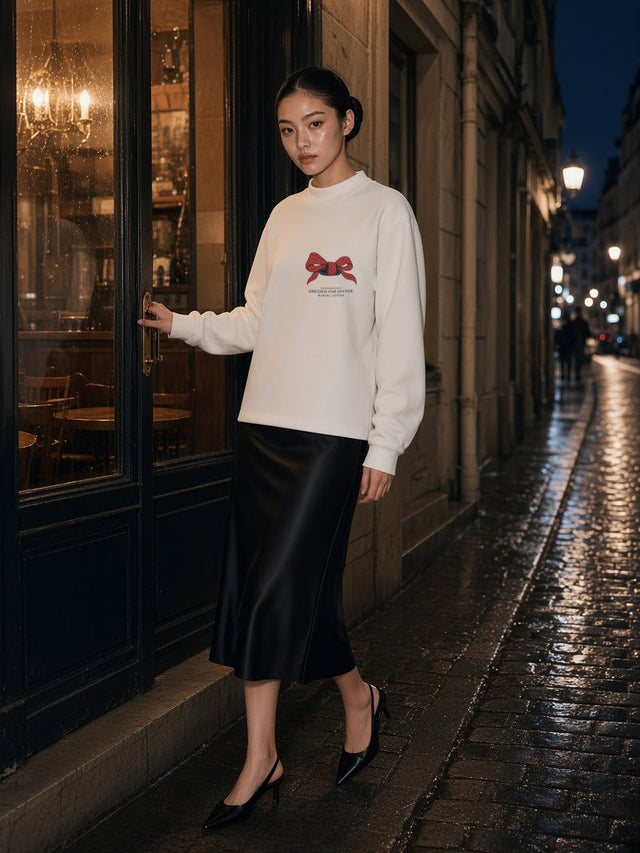 Woman in the Dressed For Dinner mock neck sweatshirt in White with the red bow print, leaving a Paris bistro on a wet cobbled street at night