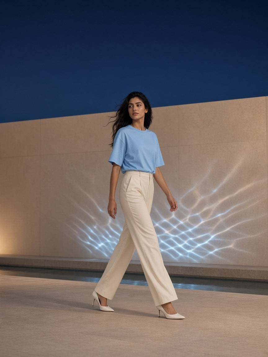 Woman in the Marcel Afters Drop Shoulder Cotton T-Shirt in Blue with ivory trousers and white heels, walking past pool-light reflections on a stone wall at night