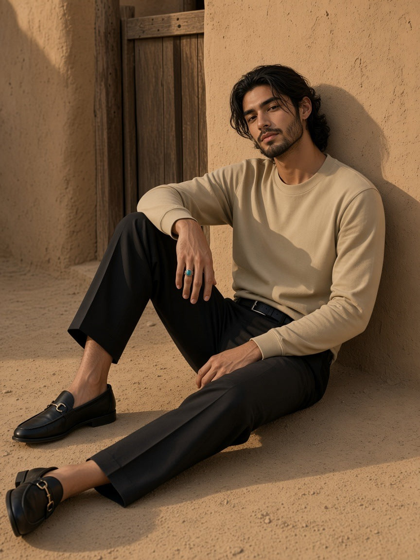 Man in the Essential Cotton Long Sleeve T-Shirt in Khaki tucked into black pleated trousers with horsebit loafers, seated against an adobe wall in a Marfa courtyard at golden hour