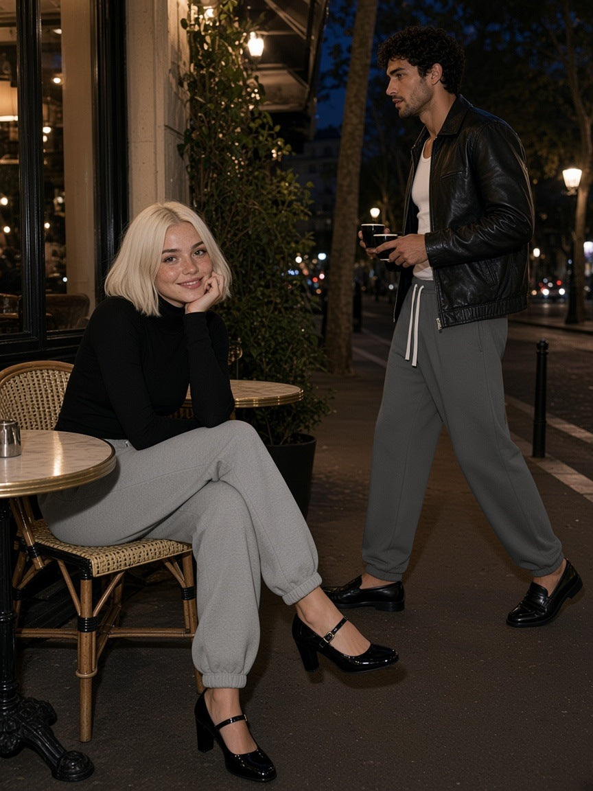 Two people in the Essential Fleece Sweatpants in Cement Gray and Carbon Gray, one at a café table and one walking past with coffee, on a Paris terrace at night