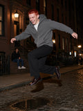 Man in the Essential Fleece Zip-Through Hoodie in Medium Gray with black trousers and loafers, leaping a puddle in Brooklyn Heights at night