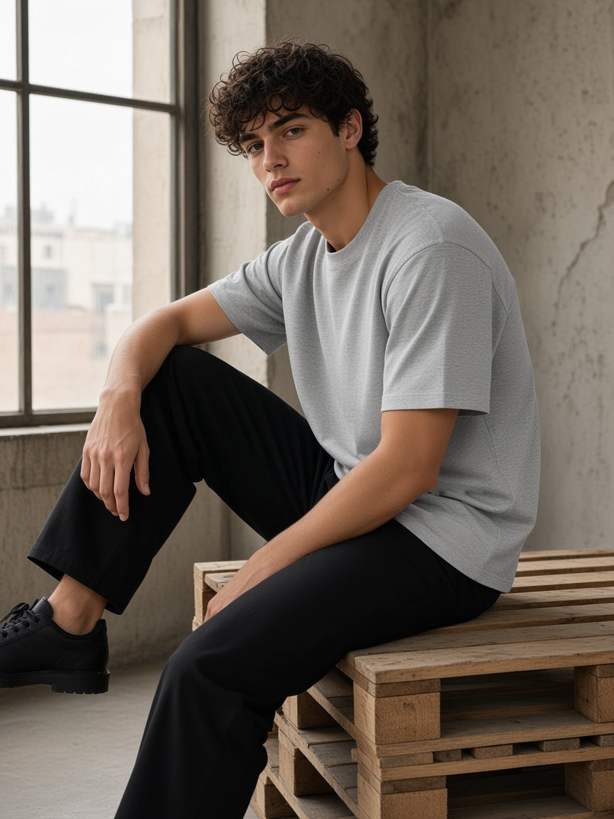 Marcel Afters Essential Heavyweight Cotton T-Shirt in Flower Gray, worn by a man, seated on a timber pallet by a tall window in a grey tee and black trousers