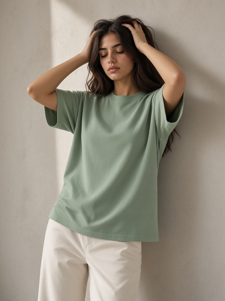 Marcel Afters Essential Heavyweight Cotton T-Shirt in Light Green, worn by a woman, leaning on a concrete wall in a sage green tee and cream trousers