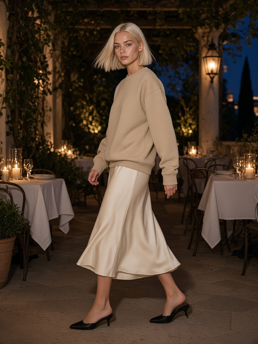 Woman in the Essential Heavyweight Crewneck Sweatshirt in Camel with an ivory silk midi skirt and kitten-heel mules, turning on a candlelit restaurant terrace at night
