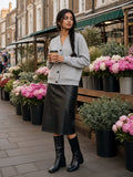 Woman in the Essential Heavyweight Sweat Cardigan in Flower Gray buttoned over a black leather midi with square-toe knee boots, coffee in hand at Columbia Road flower market, London on a Sunday morning