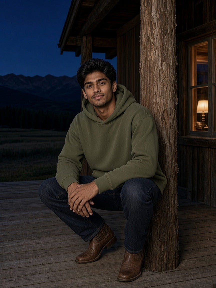 Man in an olive essential oversized hoodie and dark jeans crouched on a chalet porch at blue hour