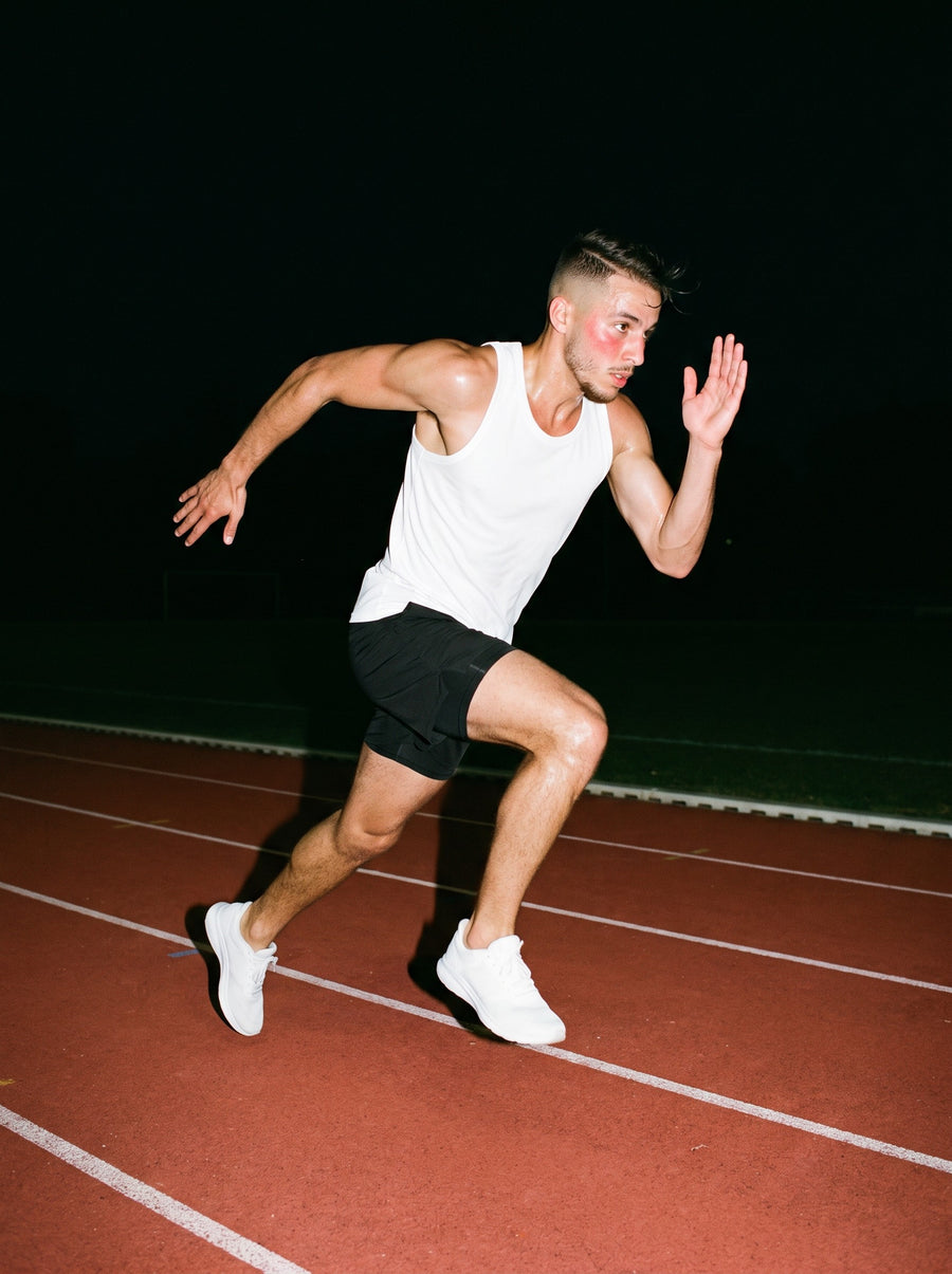 Man in the Essential Performance Tank Top in White, sprinting down a running track at night