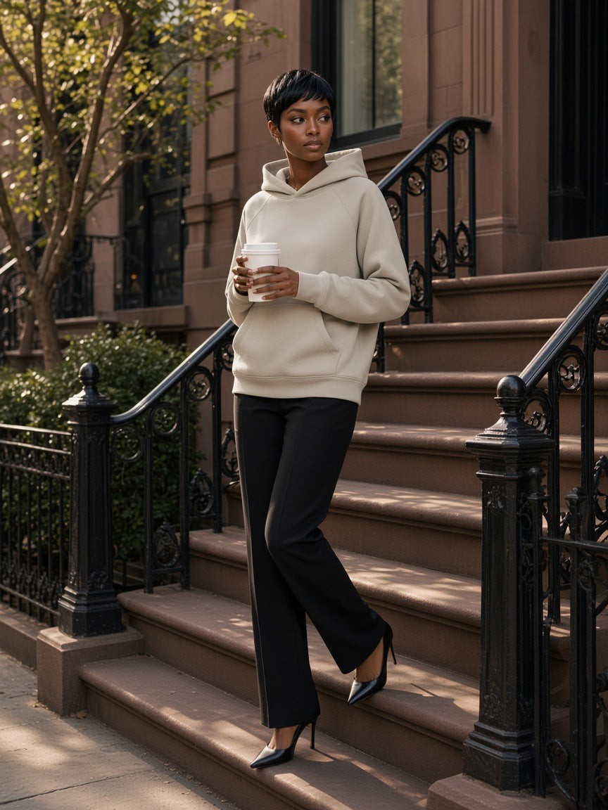 Woman in the Essential Raglan Sleeve Fleece Hoodie in Oat Gray with black trousers and heels, holding a coffee on a West Village stoop in New York in the morning