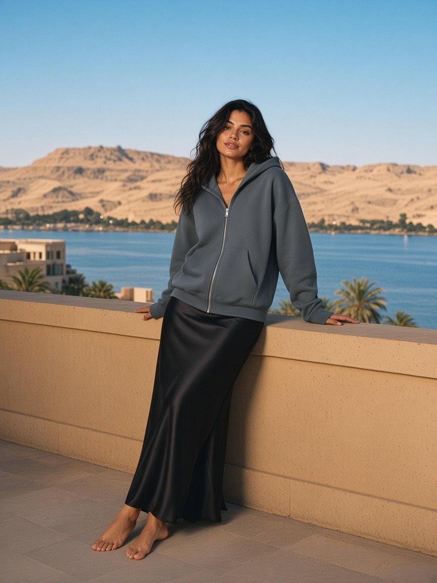 Woman in the fleeced full-zip boxy hoodie in Charcoal Gray with a black silk slip skirt, on a Nile-side hotel terrace in Luxor in the morning