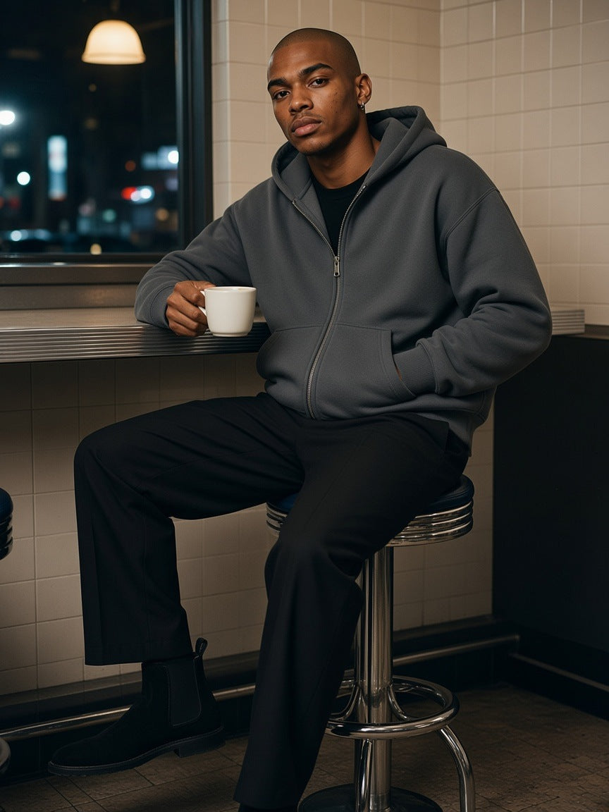 Man in the fleeced full-zip boxy hoodie in Charcoal Gray with black pleated trousers, seated at a New York diner counter at night