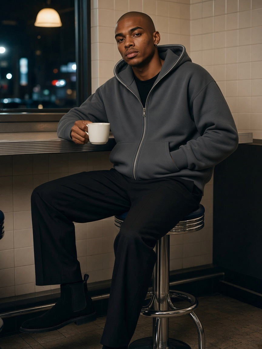 Man in the fleeced full-zip boxy hoodie in Charcoal Gray with black pleated trousers, seated at a New York diner counter at night