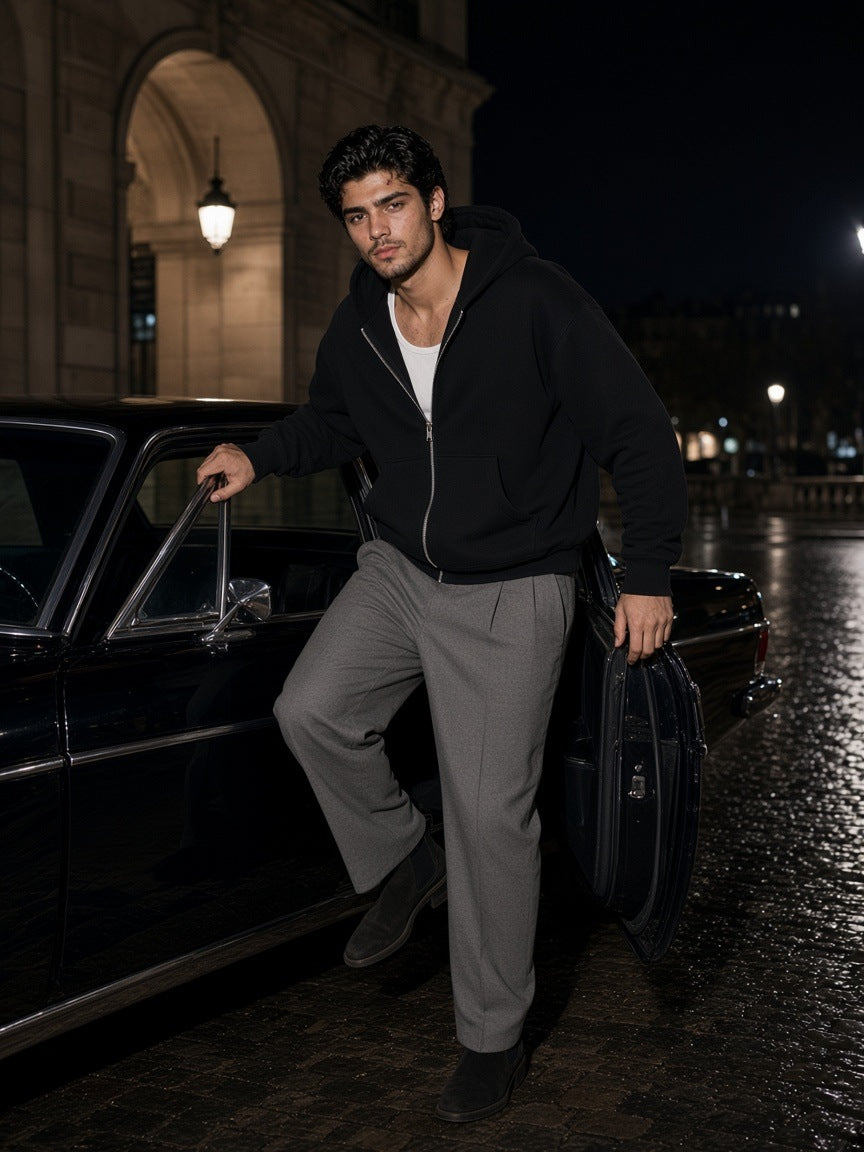 Man in the Marcel Afters fleeced full-zip boxy hoodie in Black over a white tank with grey tailored trousers, in a pale-oak hotel suite in St. Moritz at night