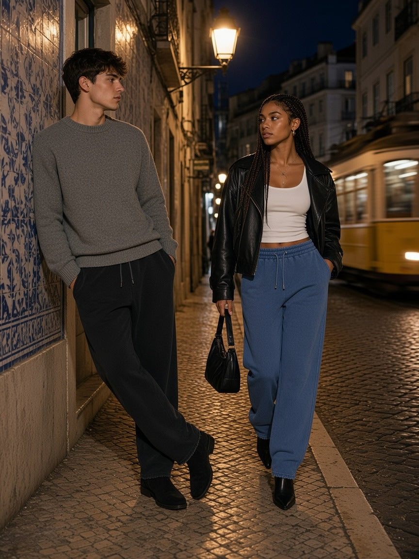 Two people in the Frayed Enzyme Washed Fleece Sweatpants in Black and Blue, one leaning on a tiled wall and one stepping off the kerb, at a Lisbon tram stop at night