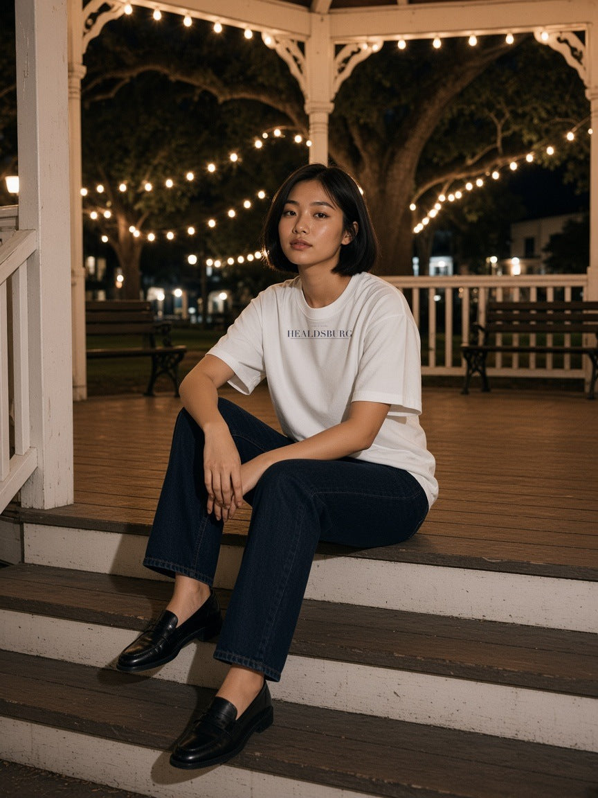 Woman in the Healdsburg Heavyweight Drop Shoulder T-Shirt in White with the navy Healdsburg city plate, leaning on a vineyard farmhouse porch post in Healdsburg at dusk