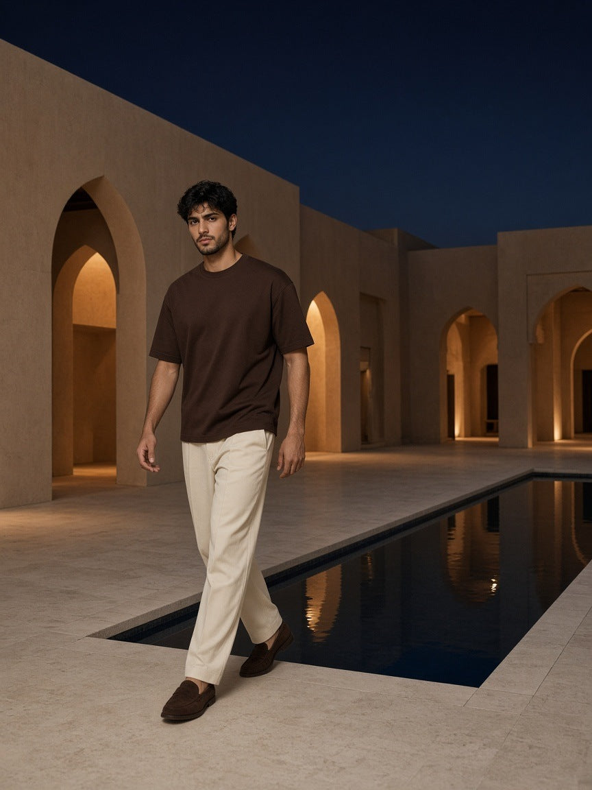 Man in the Marcel Afters Heavyweight Drop Shoulder Cotton T-Shirt in Coffee with ecru trousers and suede loafers, walking past a lit arcade pool at night