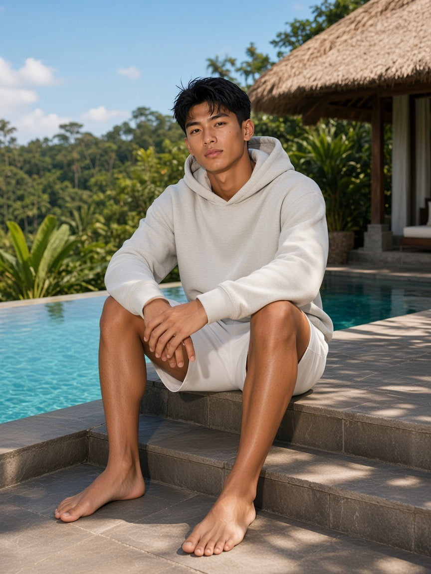 Man in the Heavyweight Pullover Hoodie in Oat Gray with white shorts, barefoot by a villa pool in Bali by day