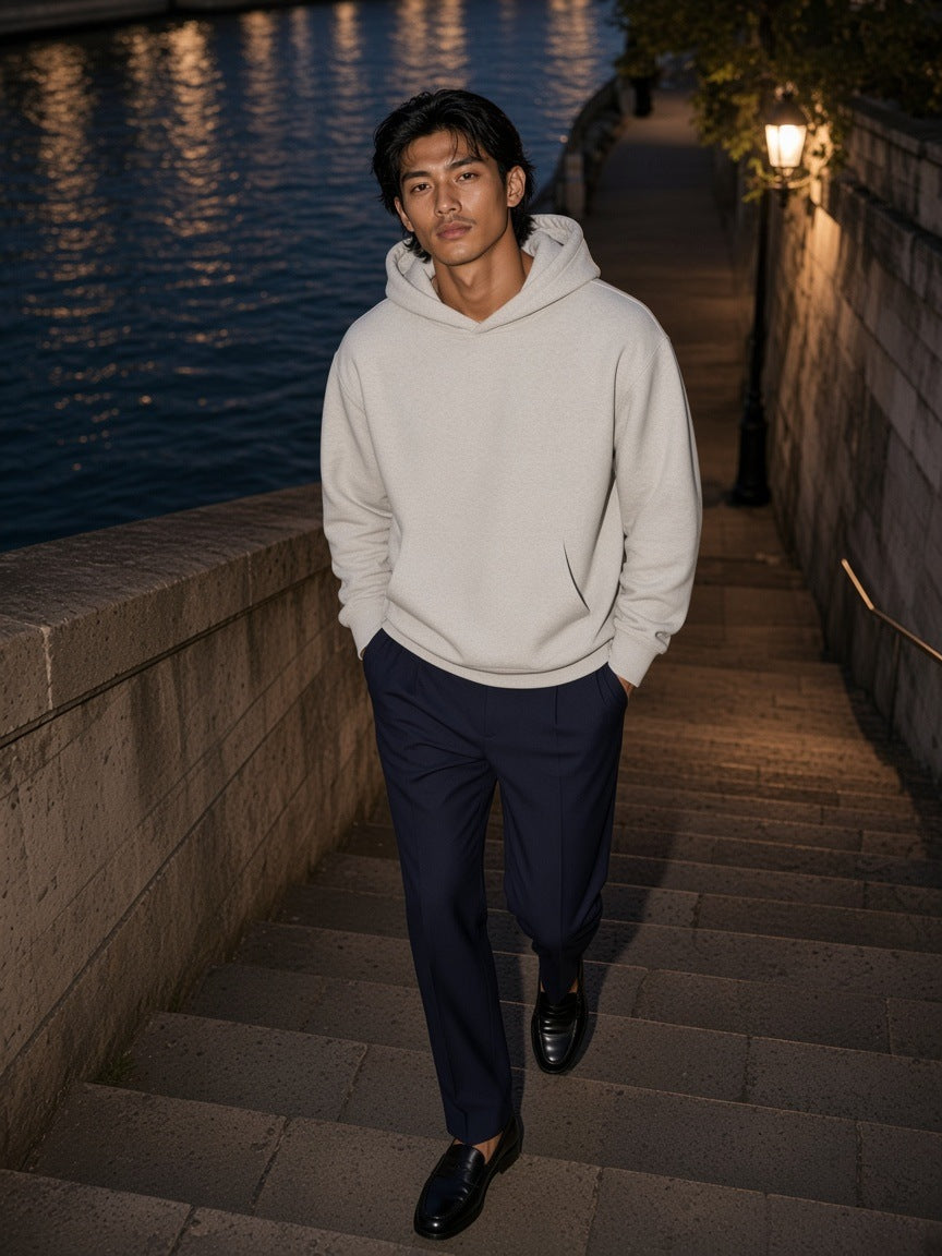 Man in the Heavyweight Pullover Hoodie in Oat Gray with navy pleated trousers and penny loafers, walking up the Seine steps by the Pont des Arts in Paris at night
