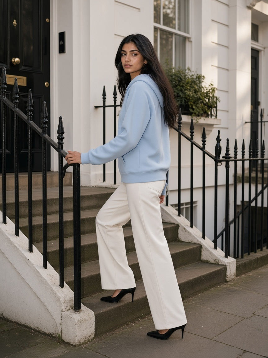 Woman in the Heavyweight Raglan Sleeve Zip Hoodie in Blue with white trousers and heels, on the front steps of a mews house in London in the morning