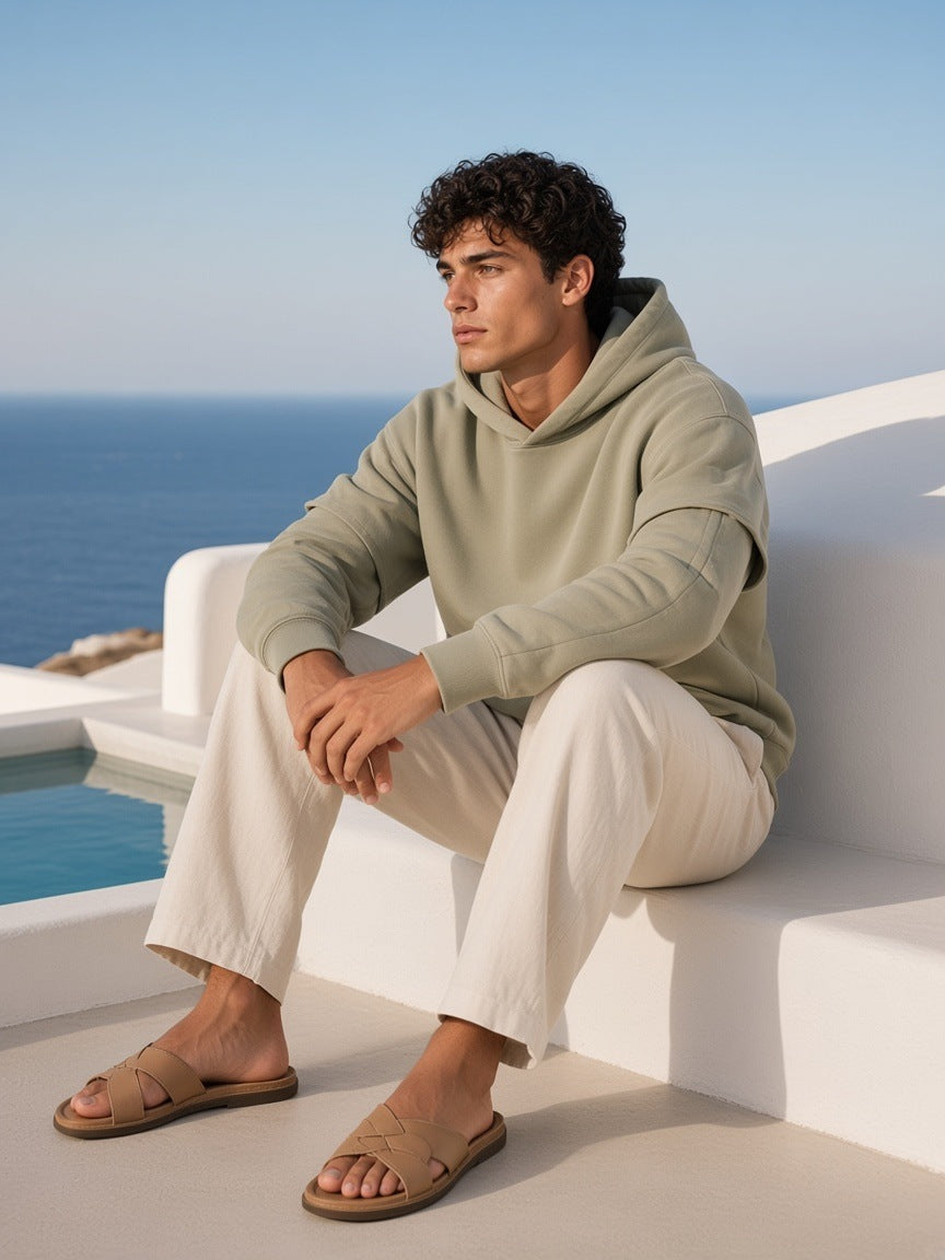 Man in the Heavyweight Ruched Sleeves Hoodie in Gray Green with linen trousers and slides, on a whitewashed cliff terrace in Mykonos in the morning