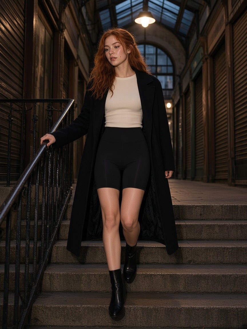 Woman in the High-Waisted Blend Biker Shorts in Black with a cream knit, long black coat and ankle boots, coming down the steps of a Paris arcade at night