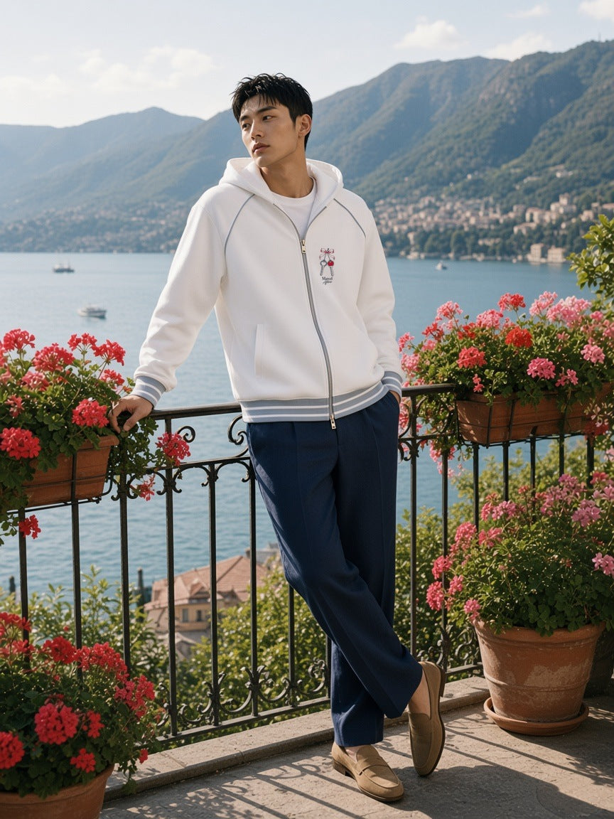 Man in the House Keys Striped Trim Zip-Up Hoodie in White leaning on a villa balcony rail above Lake Como in the morning
