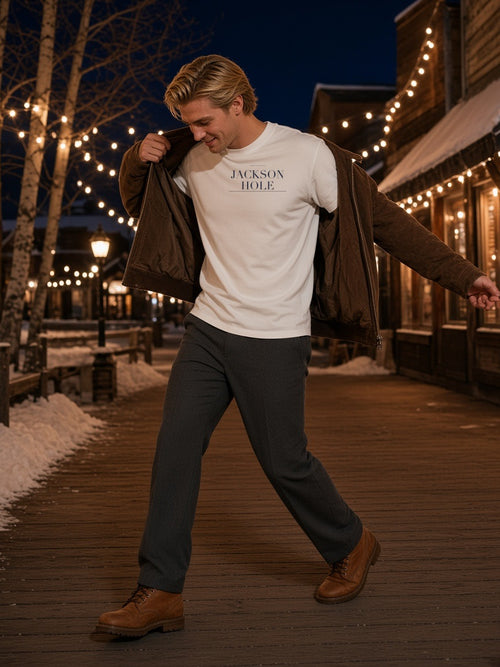 Man in the Jackson Hole Heavyweight Drop Shoulder T-Shirt in White with the navy Jackson Hole city plate, shrugging on a suede jacket on a snowy boardwalk in Jackson Hole at night