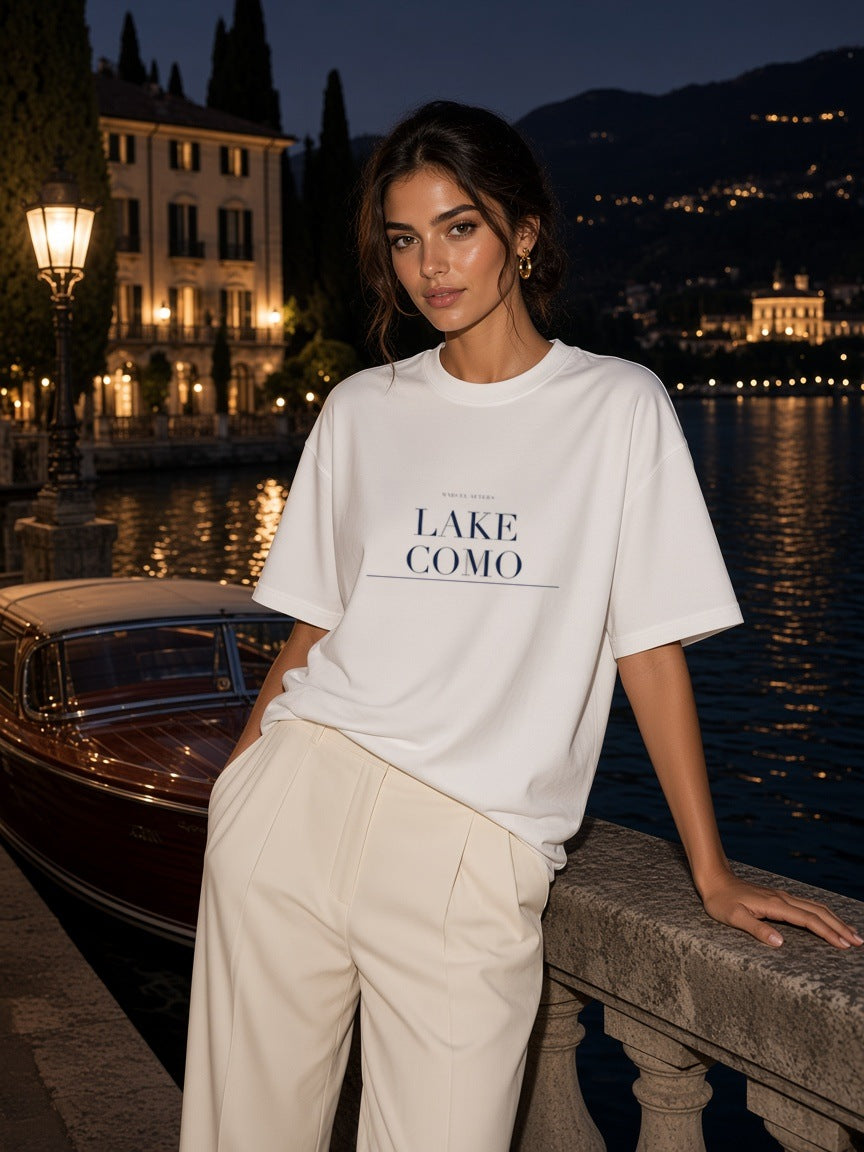 Woman in the Lake Como Heavyweight Drop Shoulder T-Shirt in White with the navy Lake Como city plate, leaning on a villa dock balustrade on Lake Como at night