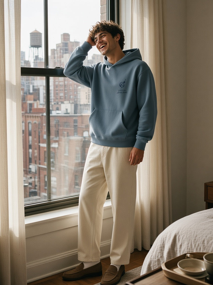Man in the Last Call Heavyweight Pullover Hoodie in Gray Blue laughing by a hotel window in New York in the morning
