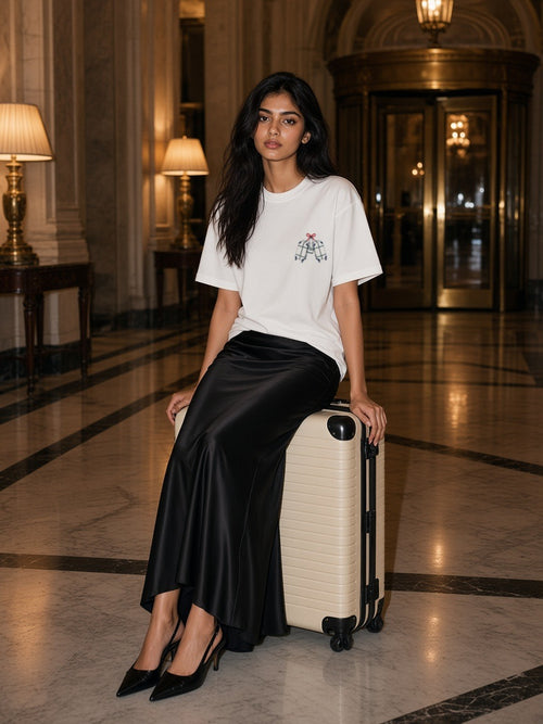 Woman in the Leaned Together tee in White seated on a suitcase in a grand hotel lobby at night