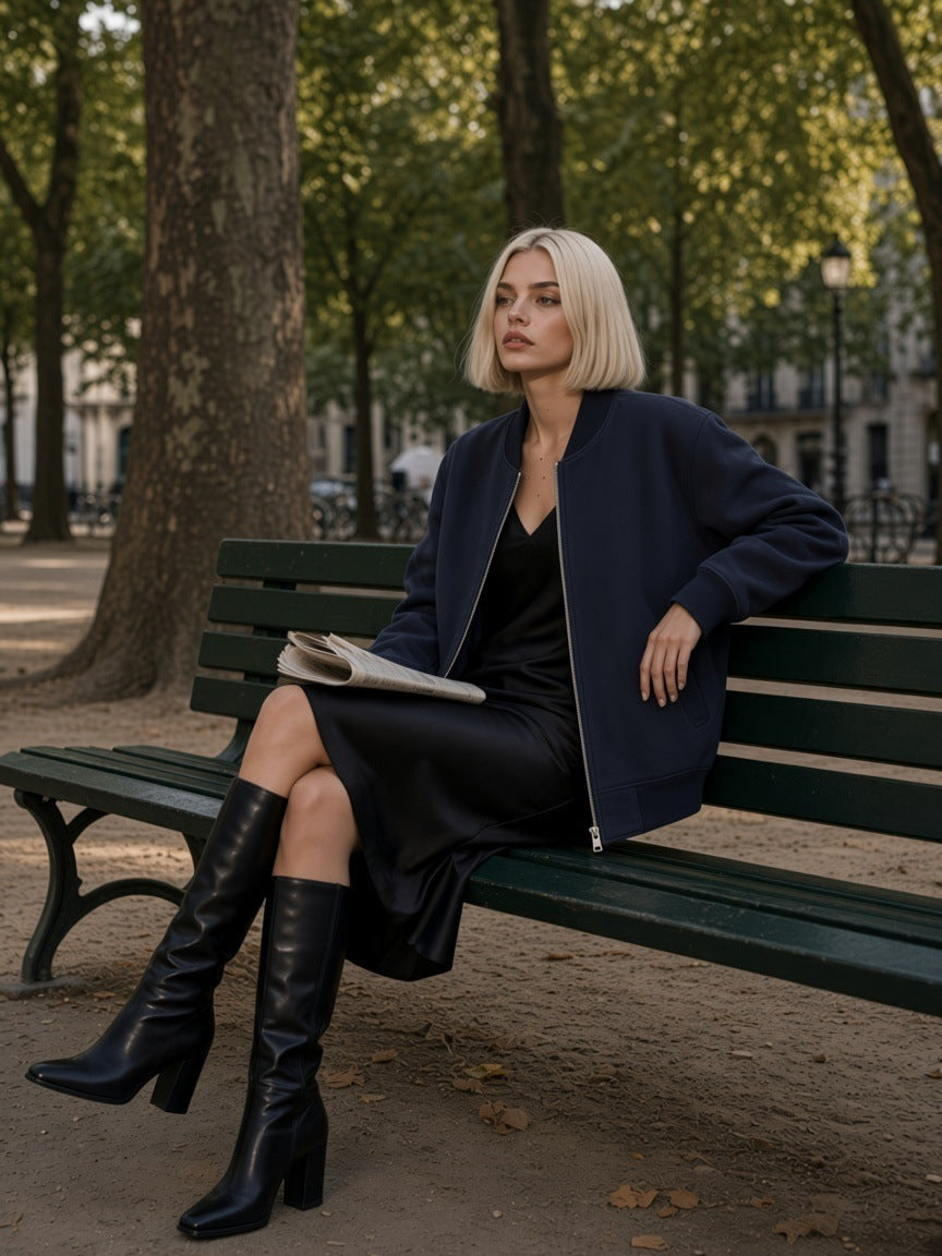 Woman in the Sun Fade Heavyweight Baseball Jacket in Navy Blue with a black satin midi and knee boots, on a bench in Place Dauphine, Paris, in the morning