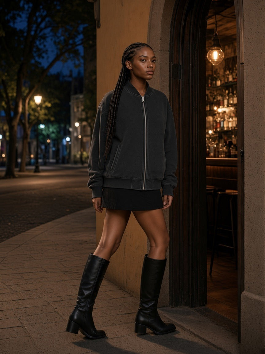 Woman in the Vintage Washed Fleece Baseball Jacket in Black with a black mini skirt and knee boots, in the lit doorway of a mezcal bar in Mexico City at night