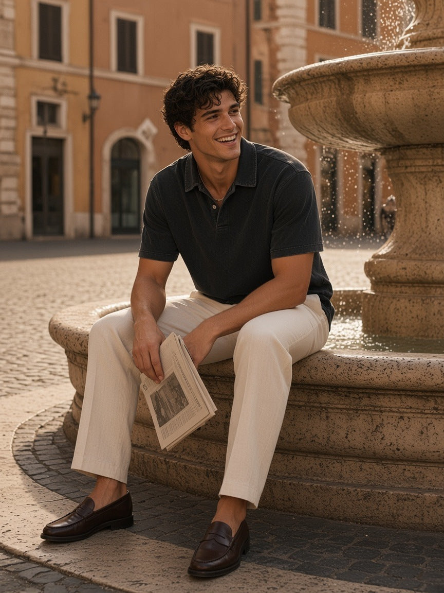 Man in the Heavyweight Snow-Washed Polo in Black with ecru trousers and penny loafers, laughing on a fountain rim in a Rome piazza in the morning