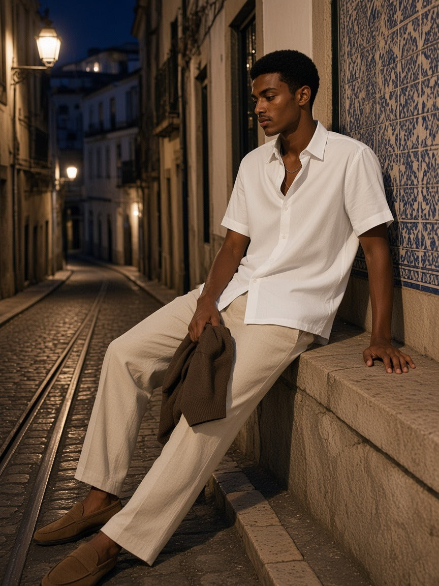 Man in the Lightweight Cotton Short Sleeve Shirt in White, worn loose with stone linen trousers, sitting by a tiled wall in Lisbon at night
